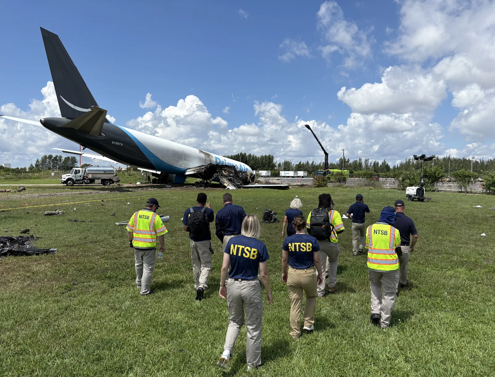 NTSB investigators walk across grass toward a damaged Amazon Prime Air Boeing 767, registration N1997A, with fire damage on the forward fuselage.